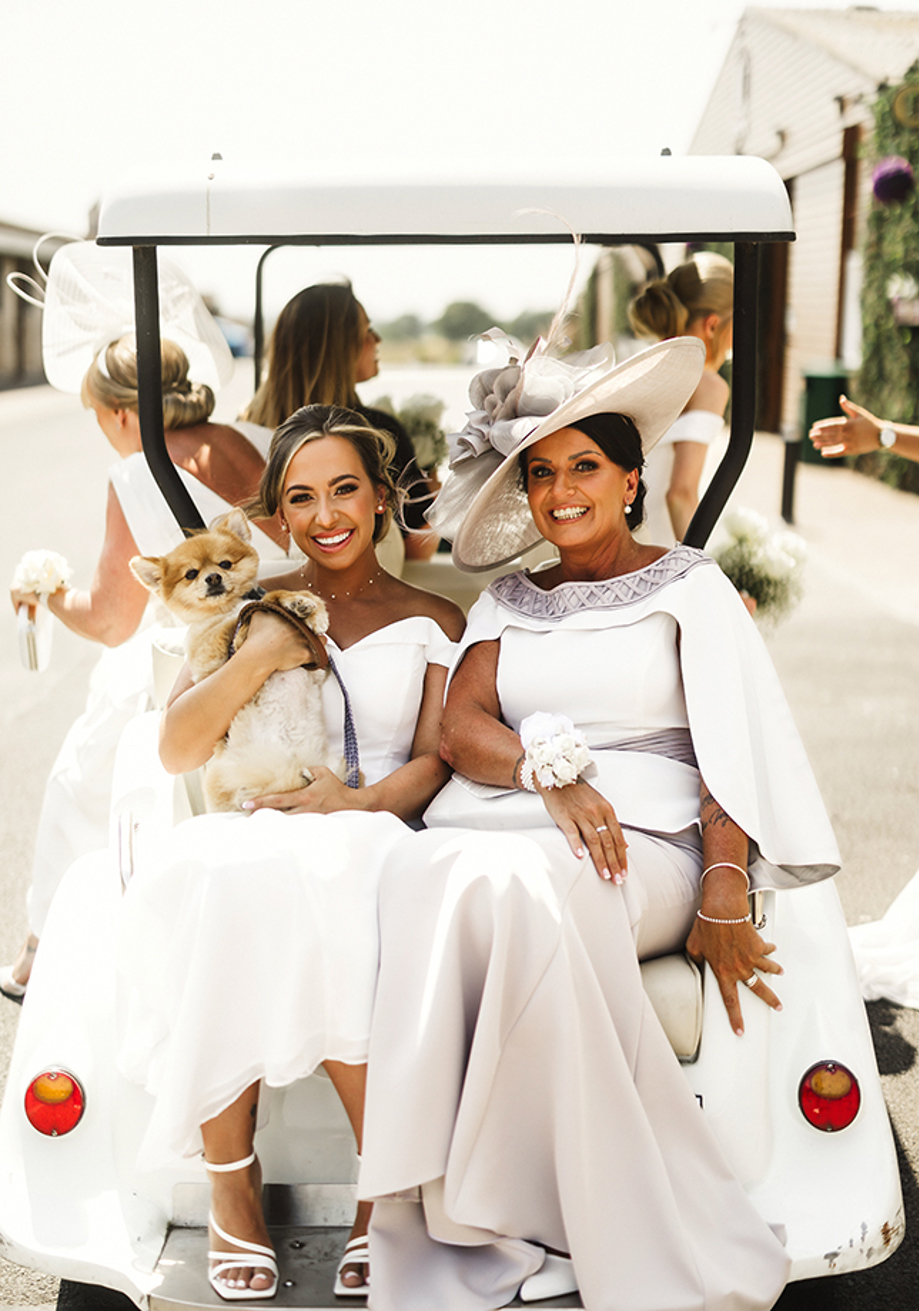 Bride and her mother take a ride on a golf cart with small dog in brides arms